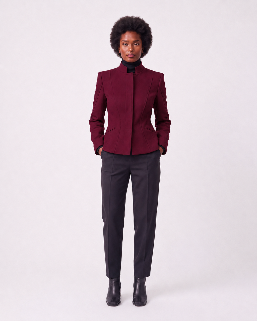 A model stands against a light gray studio background wearing a structured dark burgundy jacket with a high stand collar, paired with dark charcoal trousers and black ankle boots.