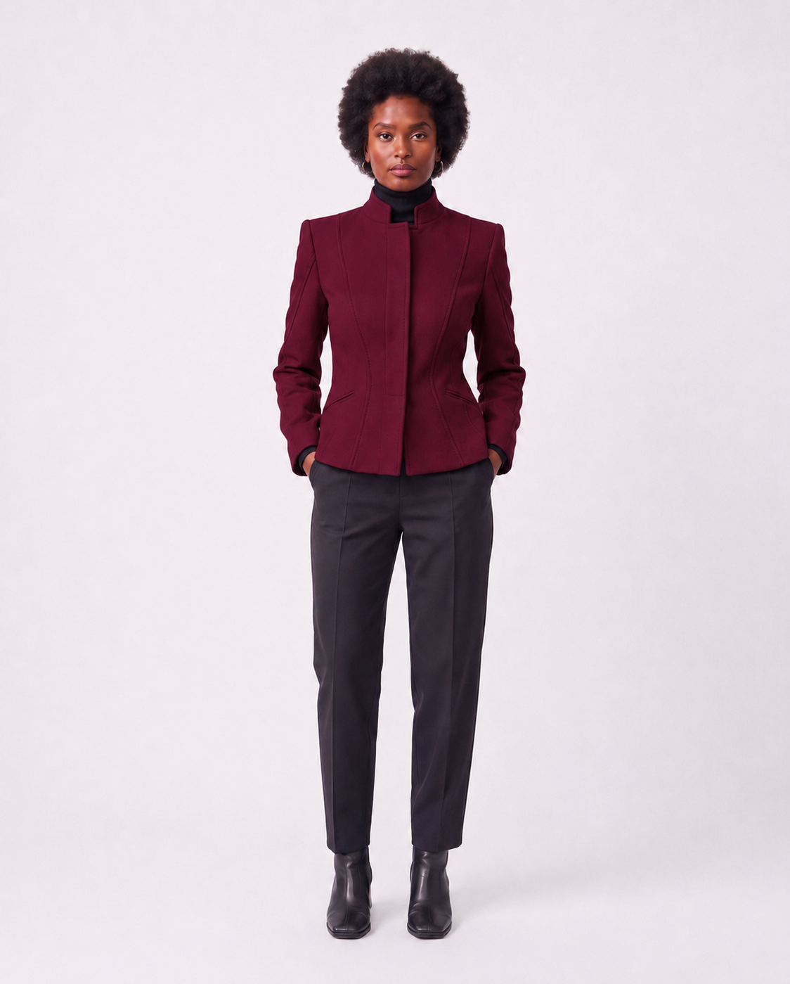 A model stands against a light gray studio background wearing a structured dark burgundy jacket with a high stand collar, paired with dark charcoal trousers and black ankle boots.