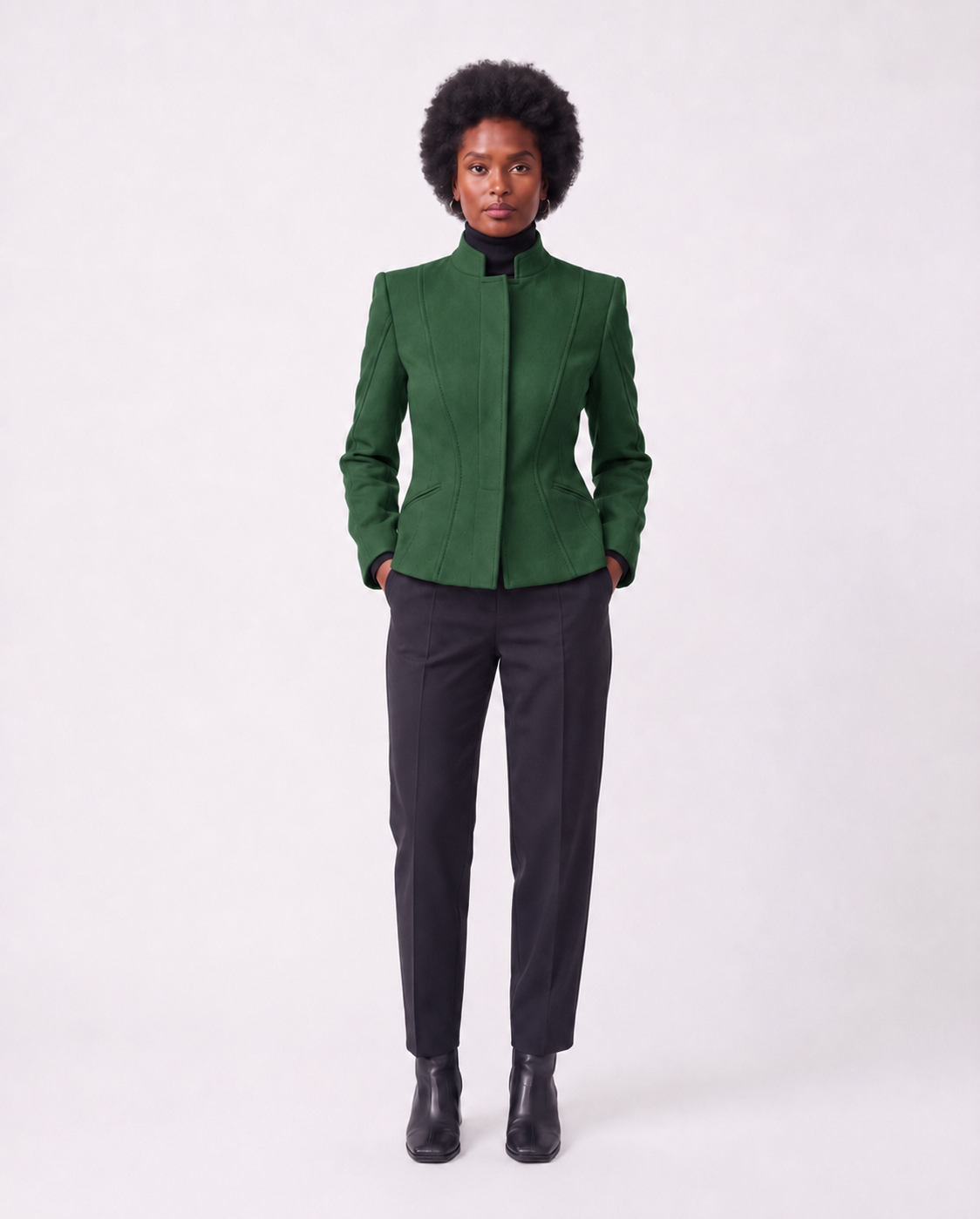 A model wearing a structured forest green cropped jacket with a stand collar, paired with dark charcoal trousers and black ankle boots, standing against a plain light gray studio background.