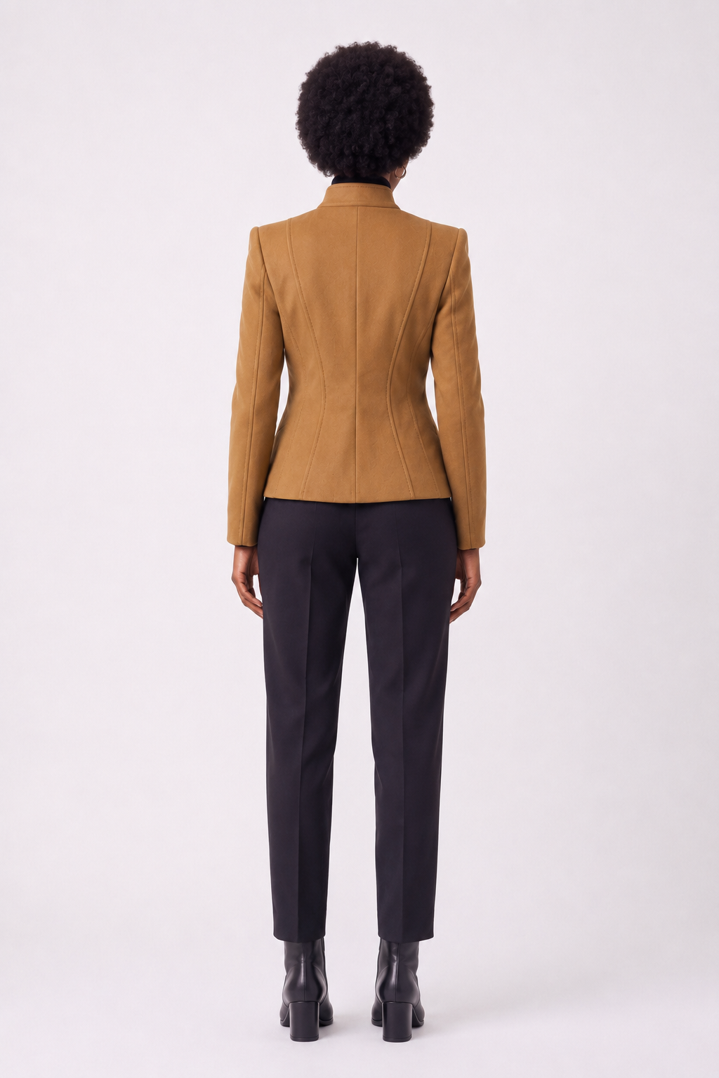 A model shown from the back wearing a camel brown structured blazer and dark charcoal trousers, standing against a light gray studio background.