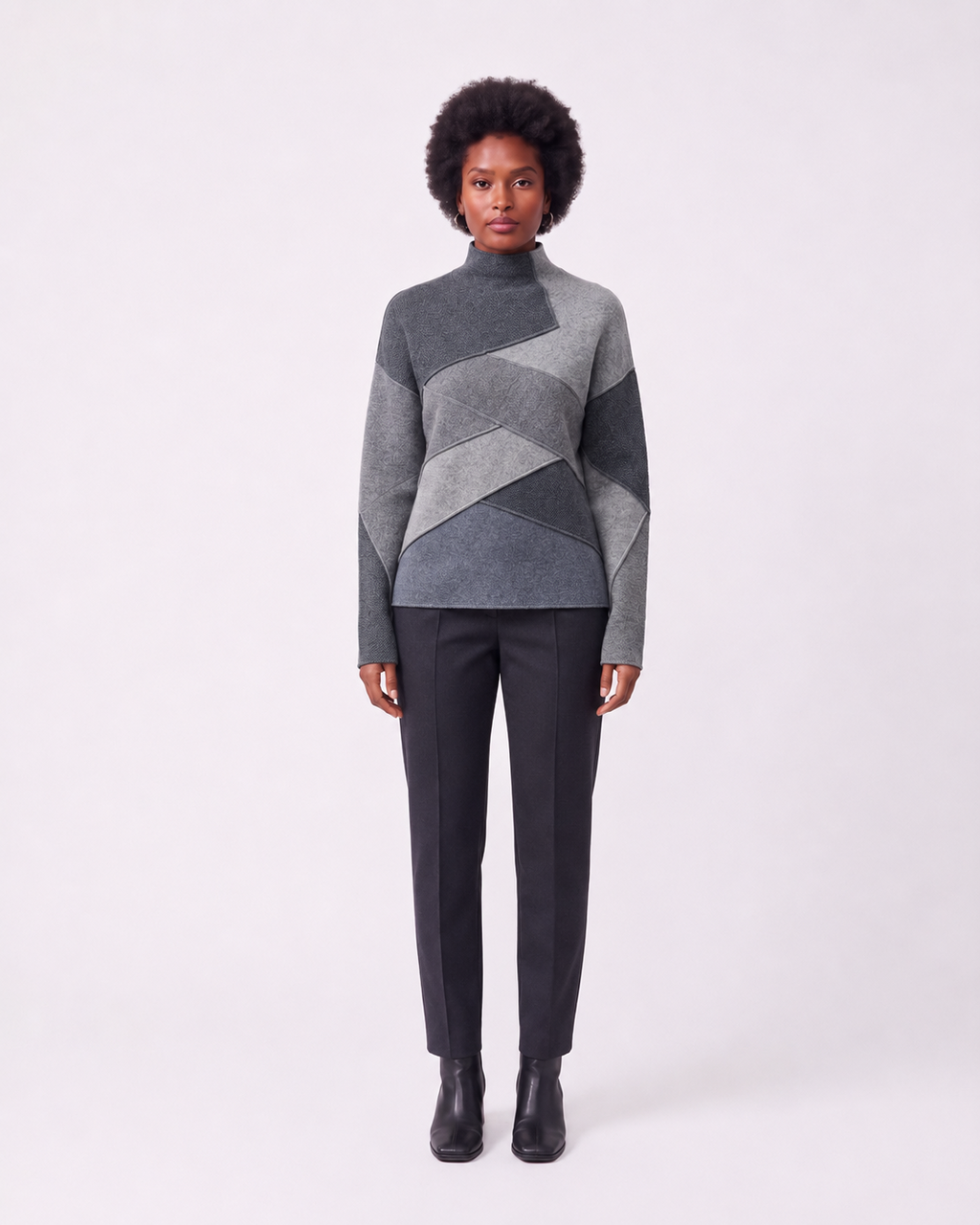 A model stands against a light gray studio background wearing a gray and charcoal geometric patchwork mock-neck sweater, dark charcoal trousers, and black ankle boots.