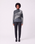 A model stands against a light gray studio background wearing a gray and charcoal geometric patchwork mock-neck sweater, dark charcoal trousers, and black ankle boots.