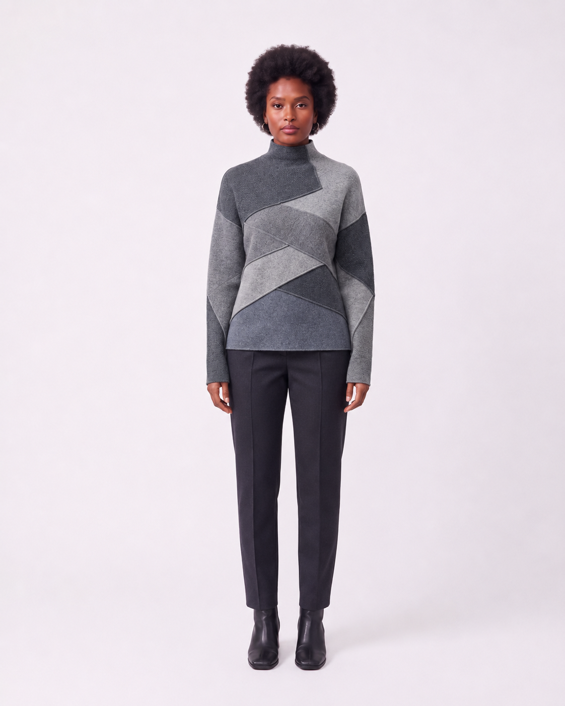 A model stands against a light gray studio background wearing a gray and charcoal geometric patchwork mock-neck sweater, dark charcoal trousers, and black ankle boots.