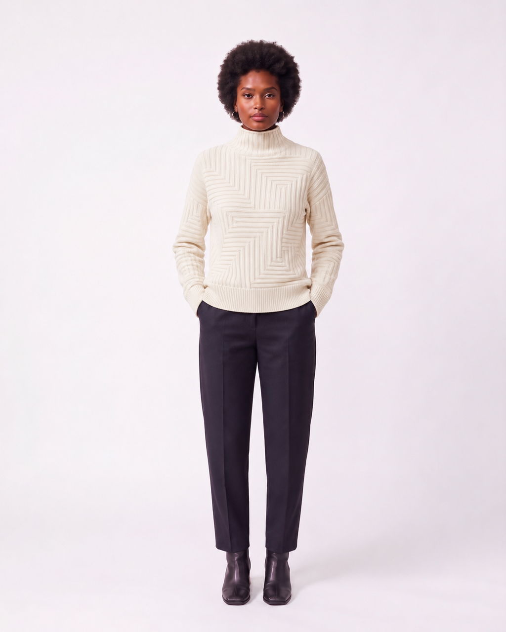 A model stands against a light gray studio background wearing a cream-colored knit mock-neck sweater with a geometric textured pattern, dark charcoal trousers, and dark brown leather ankle boots.