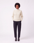 A model stands against a light gray studio background wearing a cream-colored knit mock-neck sweater with a geometric textured pattern, dark charcoal trousers, and dark brown leather ankle boots.