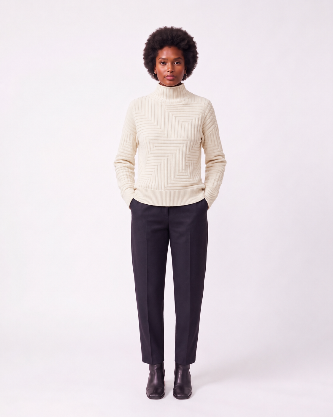 A model stands against a light gray studio background wearing a cream-colored knit mock-neck sweater with a geometric textured pattern, dark charcoal trousers, and dark brown leather ankle boots.