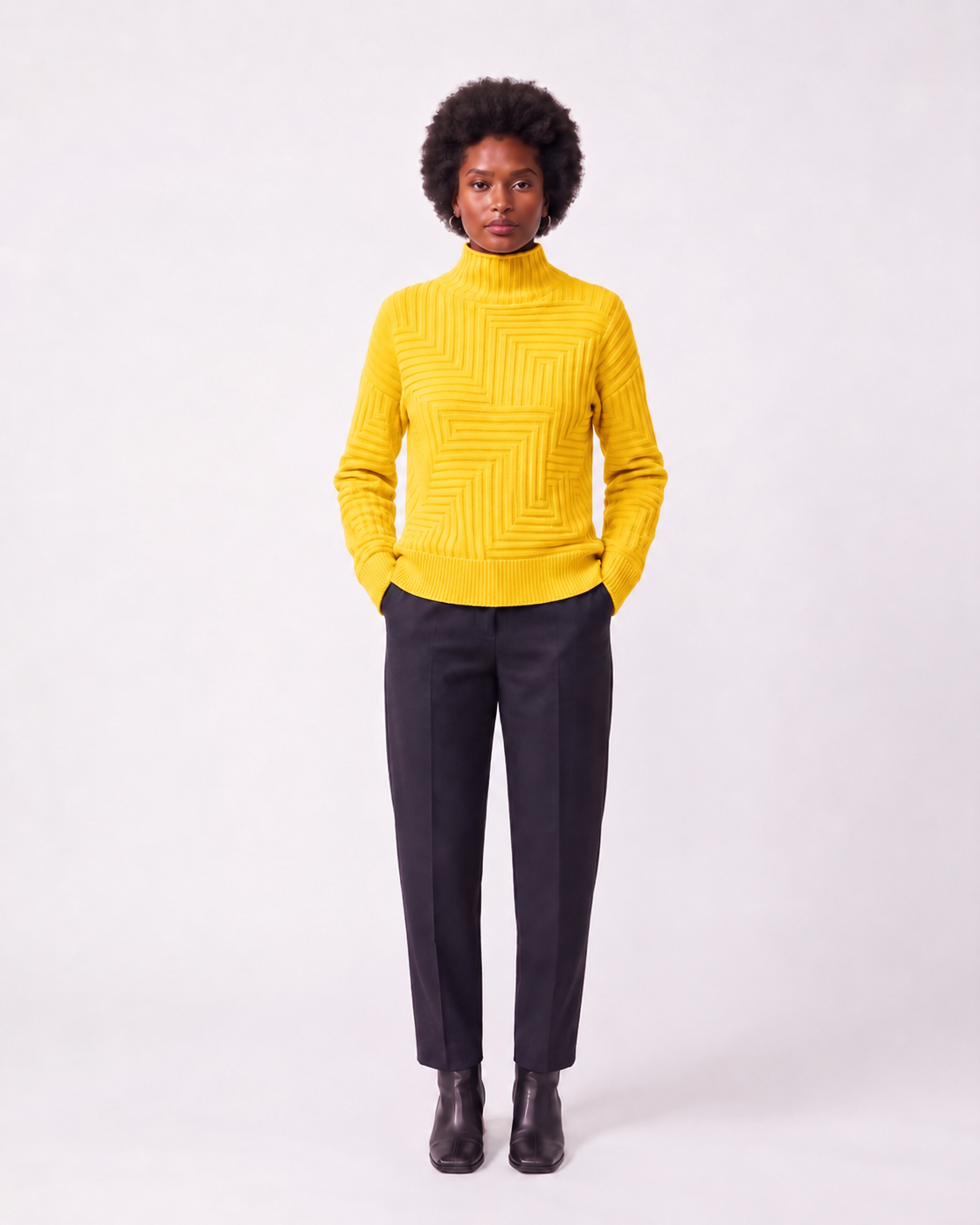 A model wearing a bright yellow ribbed knit mock neck sweater with dark charcoal trousers and black ankle boots, standing against a light gray studio background.