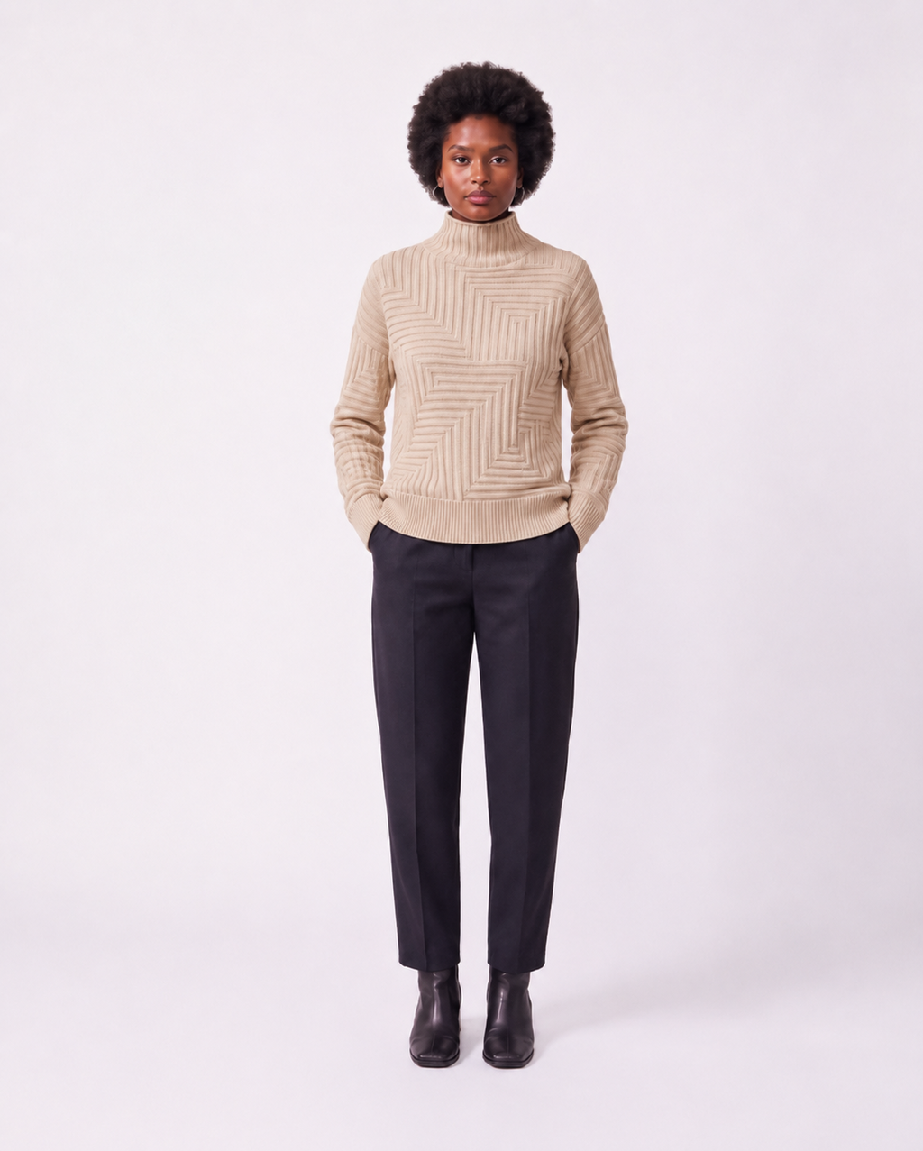 A model stands against a light gray studio background wearing a beige ribbed mock-neck sweater with a geometric knit pattern, dark charcoal trousers, and black ankle boots.