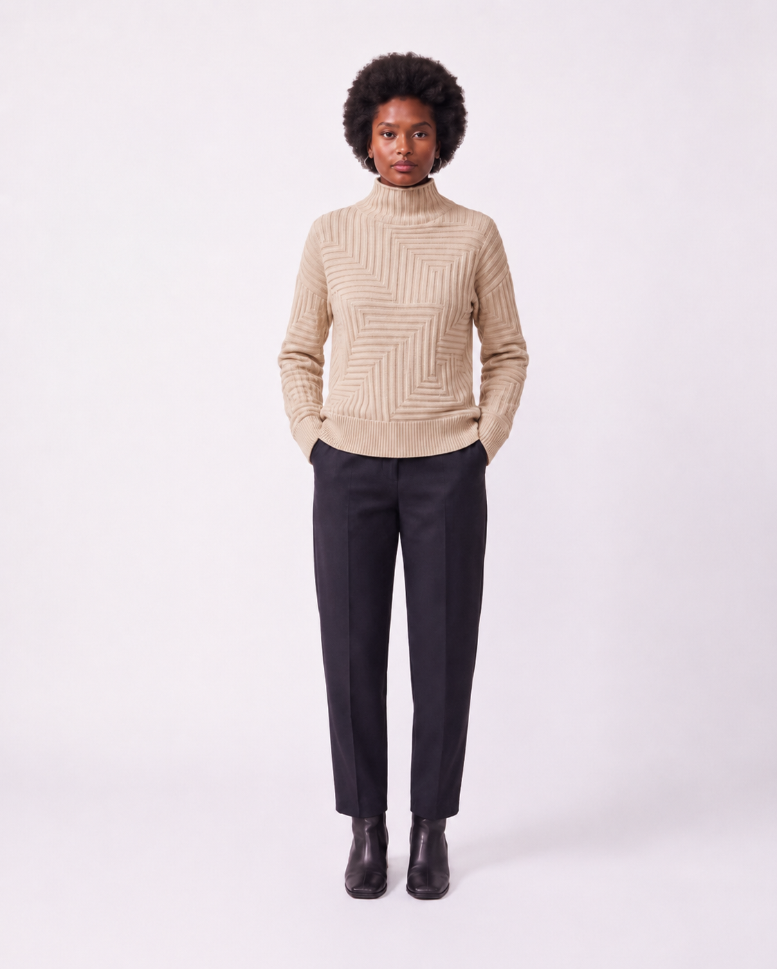 A model stands against a light gray studio background wearing a beige ribbed mock-neck sweater with a geometric knit pattern, dark charcoal trousers, and black ankle boots.