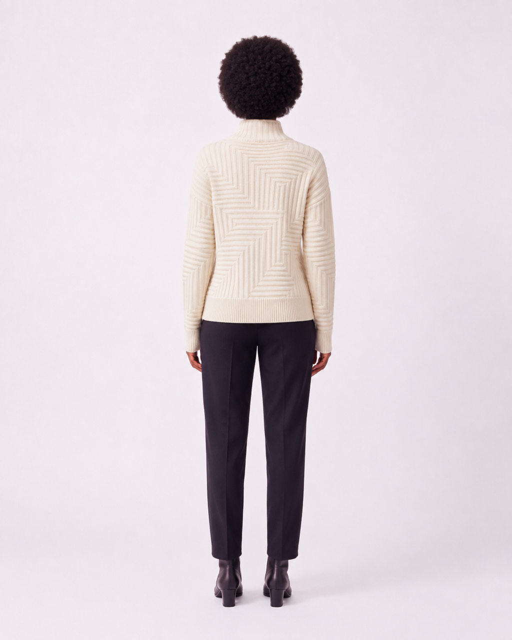 A model viewed from the back wearing a cream-colored textured knit mock-neck sweater and dark charcoal trousers, standing against a plain light gray studio background.