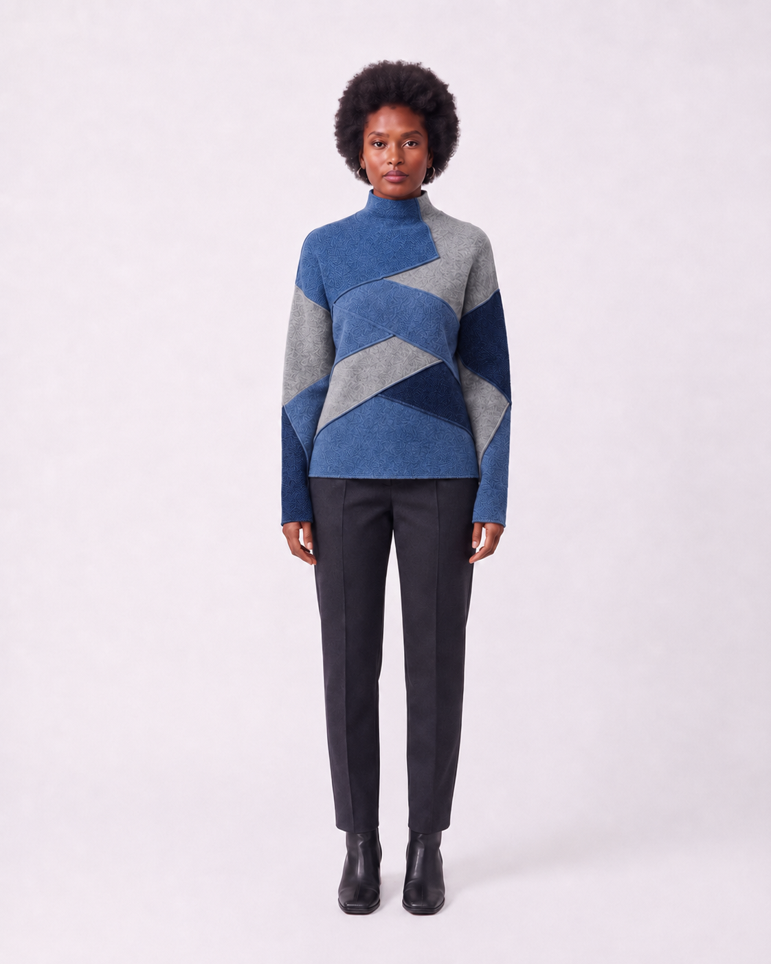 A model stands against a light gray studio background wearing a geometric color-block knit sweater with shades of blue and gray, paired with dark charcoal trousers and black ankle boots.