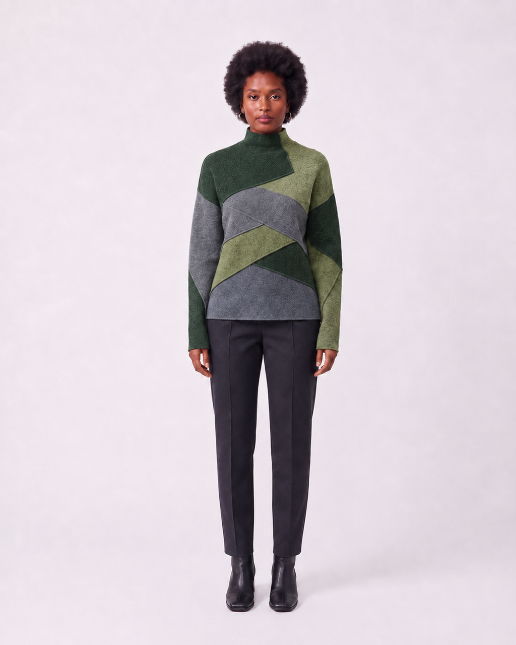 A model stands against a light gray studio background wearing a color-blocked knit mock-neck sweater with green and gray geometric panels, paired with dark charcoal trousers and black ankle boots.