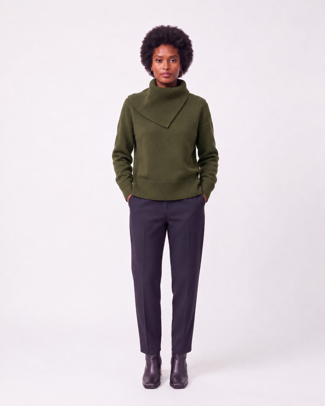 A model stands against a light gray studio background wearing an olive green knit turtleneck sweater with a notched collar, dark charcoal trousers, and dark brown leather ankle boots.