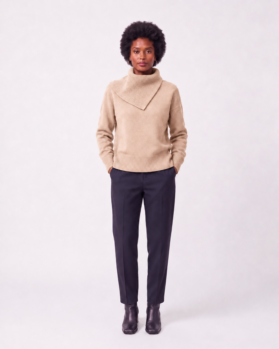 A model stands against a light gray studio background wearing a beige knit mock-neck sweater with a diagonal neckline detail, dark navy trousers, and dark brown leather ankle boots.