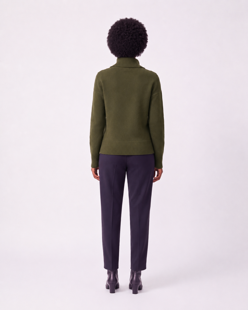 A model shown from the back wearing an olive green knit mock-neck sweater and dark charcoal trousers, standing against a plain white studio background.