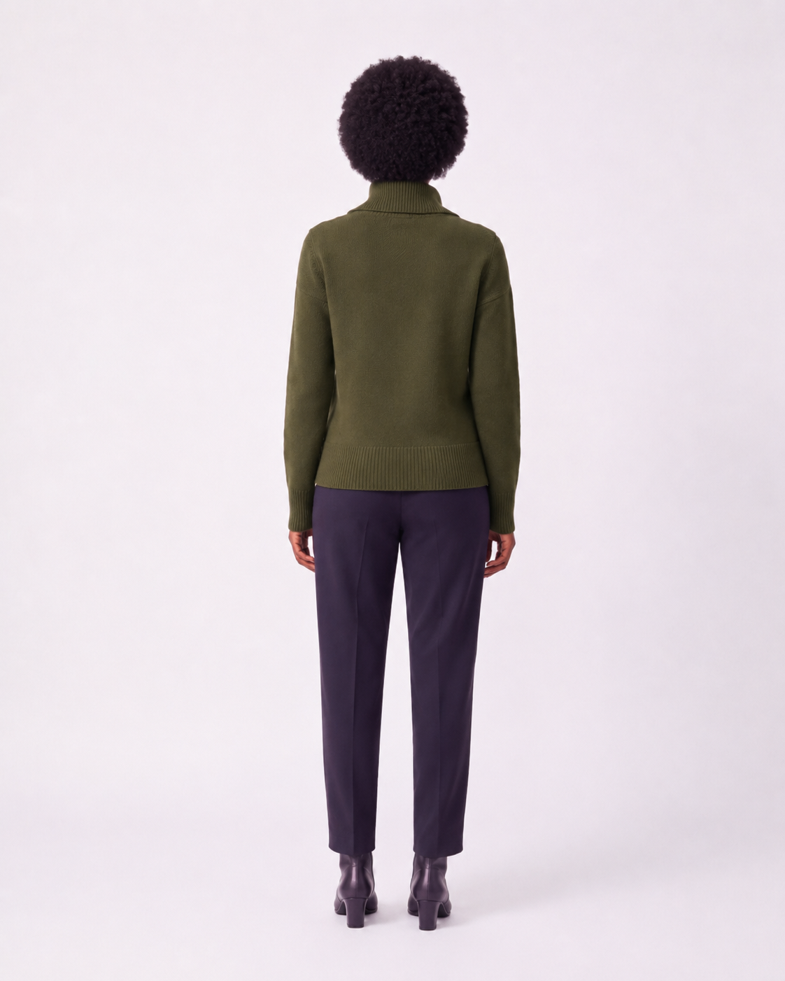 A model shown from the back wearing an olive green knit mock-neck sweater and dark charcoal trousers, standing against a plain white studio background.