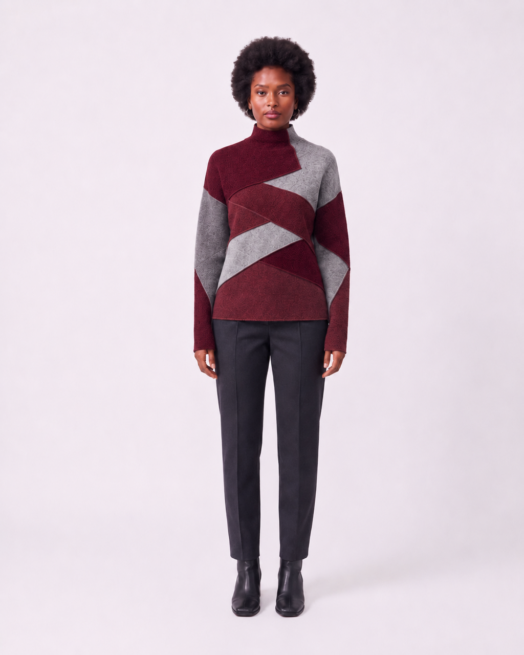 A model stands against a light gray studio background wearing a geometric color-block knit sweater with burgundy and gray panels, dark charcoal trousers, and black ankle boots.
