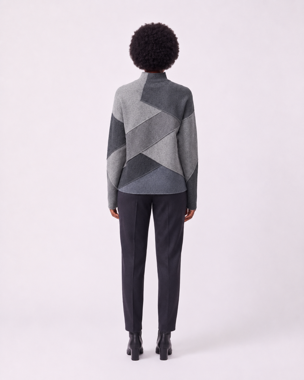 A model viewed from the back wearing a gray geometric patchwork mock-neck sweater and dark charcoal trousers against a light gray studio background.