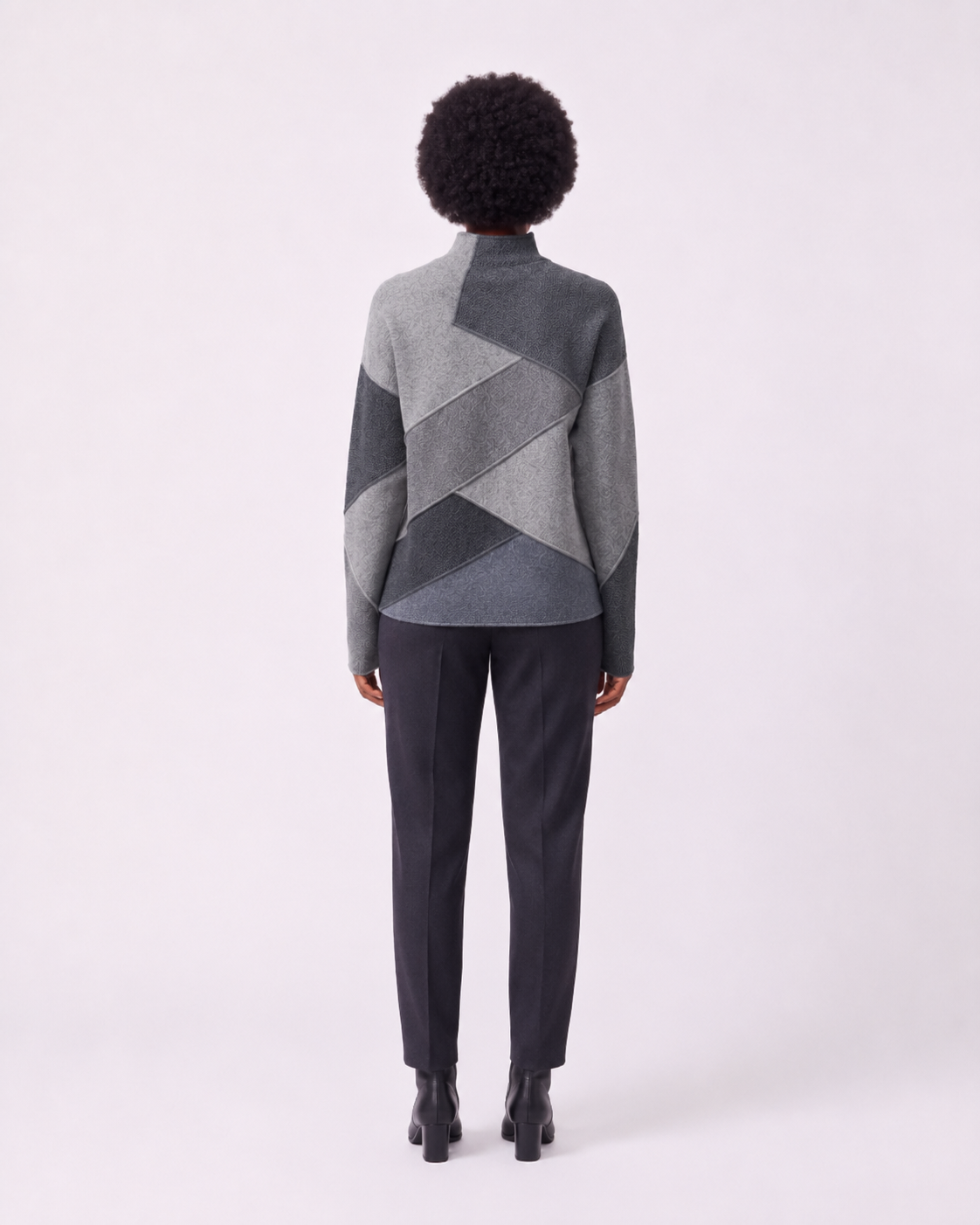 A model viewed from the back wearing a gray geometric patchwork mock-neck sweater and dark charcoal trousers against a light gray studio background.