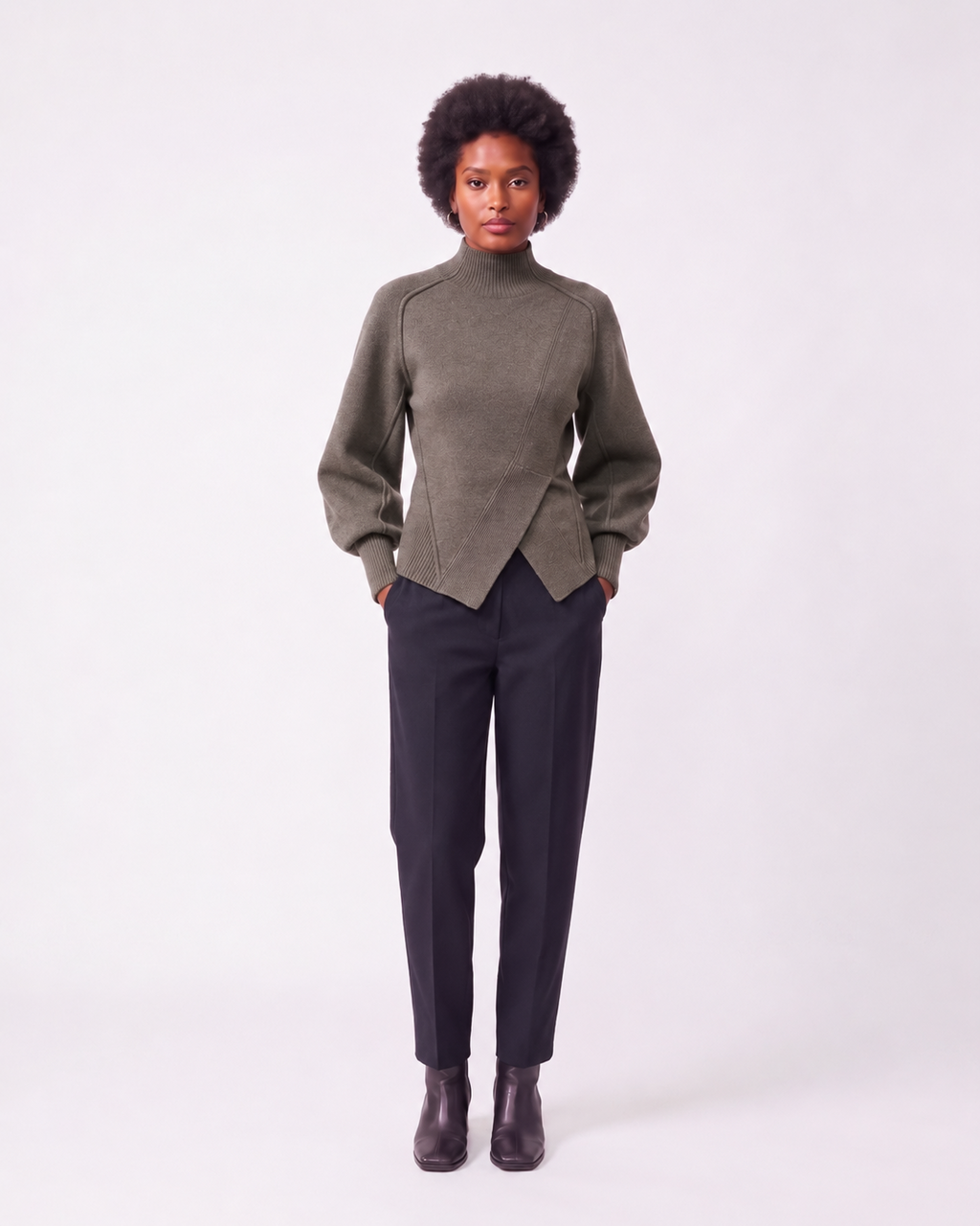 A model stands against a light gray studio background wearing a muted olive green mock-neck wrap sweater, dark charcoal trousers, and dark brown leather ankle boots.