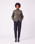 A model stands against a light gray studio background wearing a muted olive green mock-neck wrap sweater, dark charcoal trousers, and dark brown leather ankle boots.