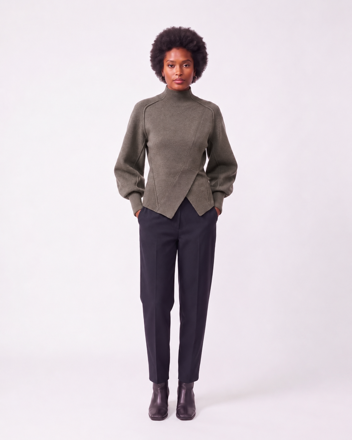 A model stands against a light gray studio background wearing a muted olive green mock-neck wrap sweater, dark charcoal trousers, and dark brown leather ankle boots.