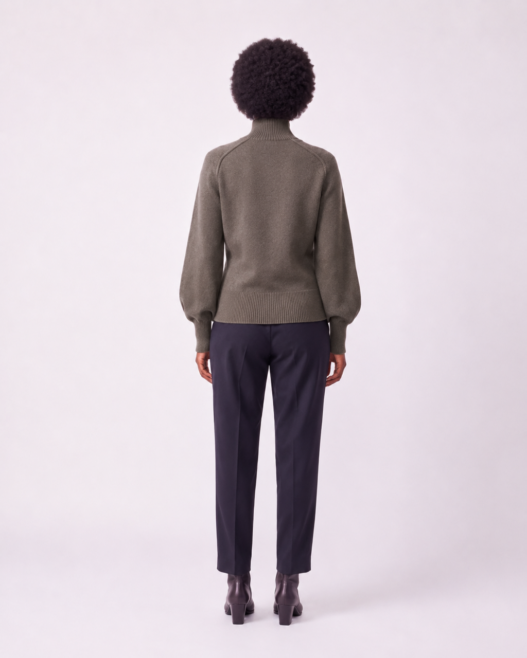 A model viewed from the back wearing an olive green knit sweater with balloon sleeves and dark navy trousers, set against a plain light gray studio background.