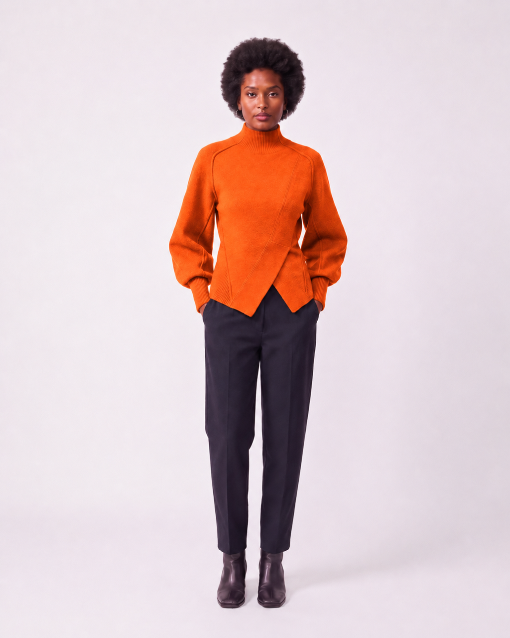 A model stands against a light gray studio background wearing a bright orange mock-neck knit sweater with long sleeves and dark charcoal trousers.